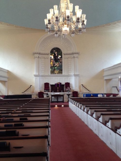 Interior of a church featuring pews, a chandelier, and a stained glass window at the front.