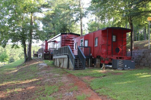 A red train caboose converted into a cabin, surrounded by trees and grass, with steps leading up to it.