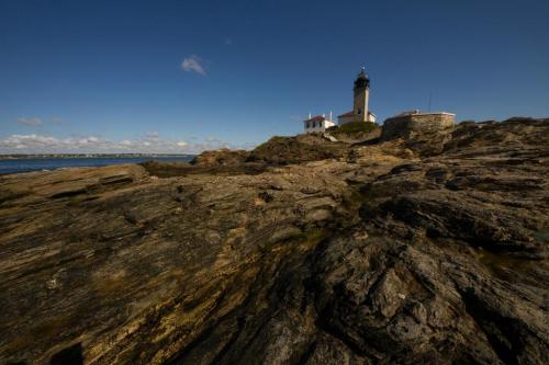A lighthouse stands on rocky terrain by the sea under a clear blue sky with scattered clouds.