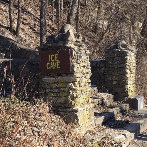 Stone pillars with a sign reading "ICE CAVE" beside a rocky path in a wooded area.