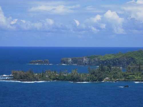 A scenic view of a coastal landscape with lush greenery, rocky cliffs, and a vibrant blue ocean under a partly cloudy sky.