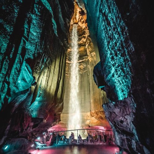 A stunning waterfall cascades down a rocky cave, illuminated by colorful lights, creating a serene atmosphere.