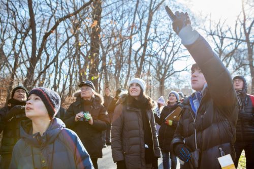 A group of people, including children, look up at the sky in a wooded area, some pointing and smiling.