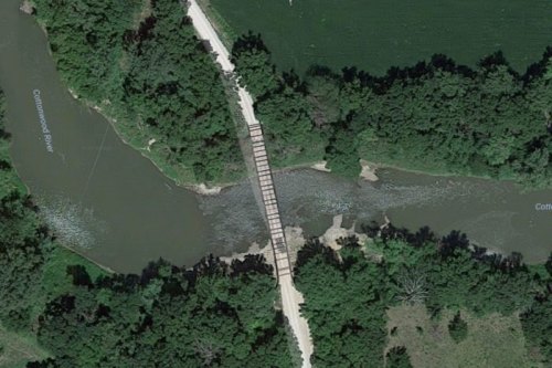 Aerial view of a wooden bridge crossing a river, surrounded by lush greenery and fields.