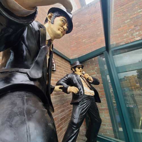 Two large statues of men in black suits and hats, posed as if performing, set against a brick wall and glass enclosure.