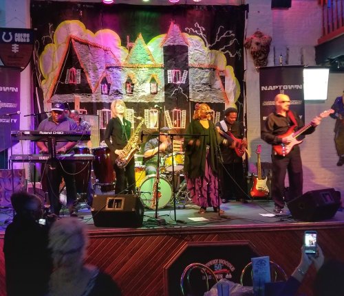 A live band performs on stage with various instruments, colorful lights, and a vibrant backdrop in a lively venue.
