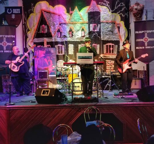 A band performs on stage with instruments, set against a colorful backdrop featuring a haunted house theme.