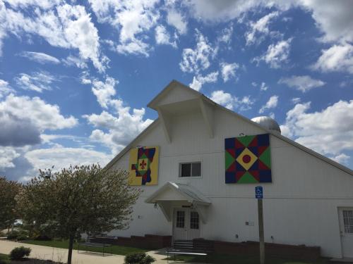 A white barn with colorful quilt patterns on its sides, under a blue sky with fluffy clouds.