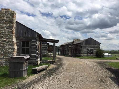 A gravel path leads between rustic wooden buildings under a cloudy sky. Green grass surrounds the area.