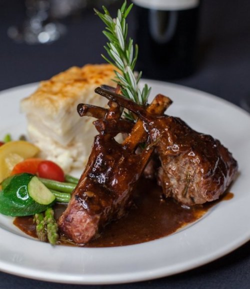 A gourmet plate featuring two lamb chops with a rich sauce, accompanied by vegetables and a creamy side dish.