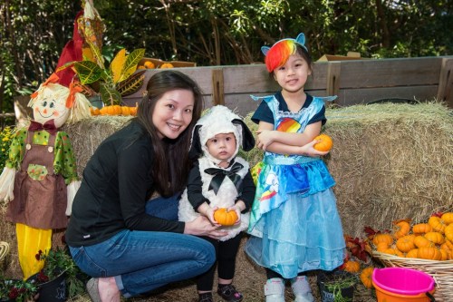 A woman and two children pose with pumpkins, one in a sheep costume and the other in a colorful dress, surrounded by autumn decor.