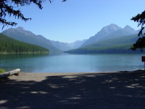 A serene lake surrounded by mountains and lush forests under a clear blue sky.