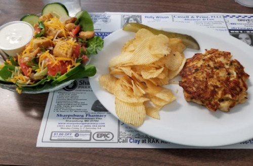 A fresh salad with lettuce, tomatoes, cheese, and croutons, served with a plate of chips and a crab cake.