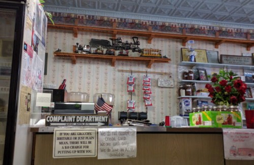 A cozy diner counter with a model train display, complaint department sign, and various jars and decorations.