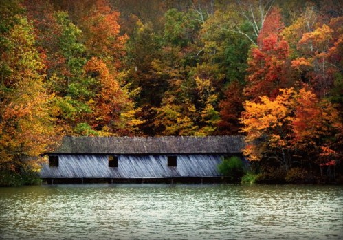 A rustic wooden structure by a calm lake, surrounded by vibrant autumn foliage in shades of orange and red.