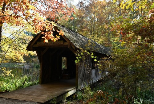 A rustic wooden covered bridge surrounded by vibrant autumn foliage and a serene pond.