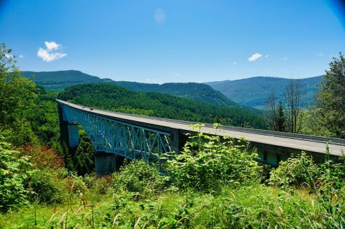A blue bridge spans a valley surrounded by lush green hills under a clear blue sky.