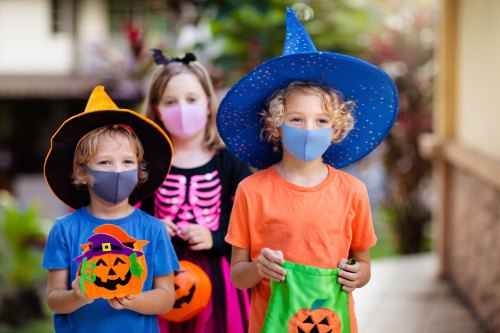 Three children in Halloween costumes wear masks, holding pumpkin bags, with a colorful outdoor background.