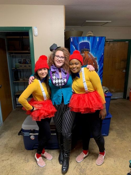 Three people pose together in a room, two in colorful costumes with red hats and tutus, and one in a stylish outfit.