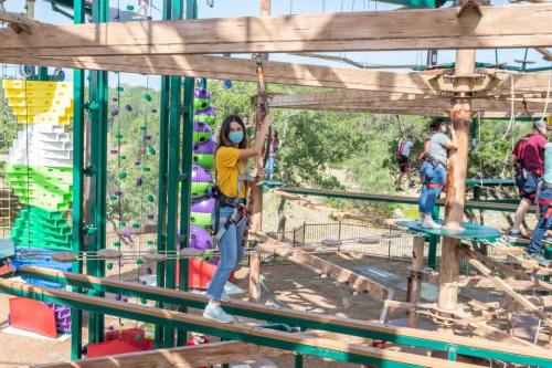A person in a yellow shirt and mask navigates a ropes course, surrounded by trees and colorful climbing structures.