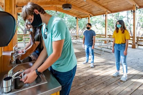 Four people wearing masks wash their hands at outdoor sinks in a wooden pavilion.