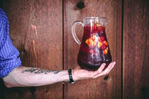 A person holds a glass pitcher filled with dark red beverage and ice, set against a wooden background.
