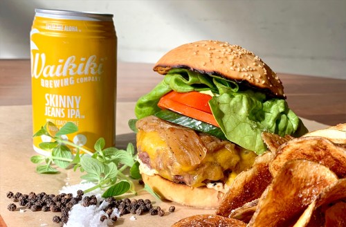 A delicious burger with lettuce, tomato, and cheese, served with potato chips and a can of Skinny Jeans IPA.