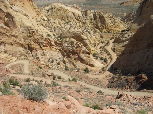 Winding dirt road through rocky terrain, surrounded by steep cliffs and sparse vegetation in a desert landscape.