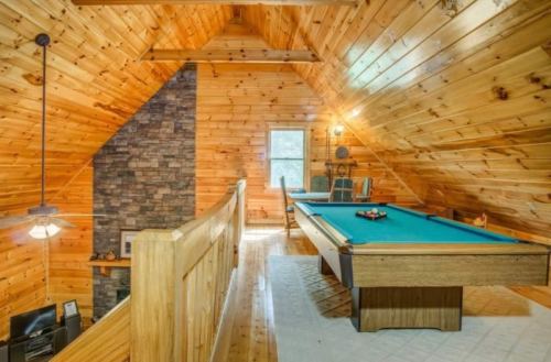 Cozy wooden loft with a pool table, stone wall, and large windows, featuring warm, rustic decor.