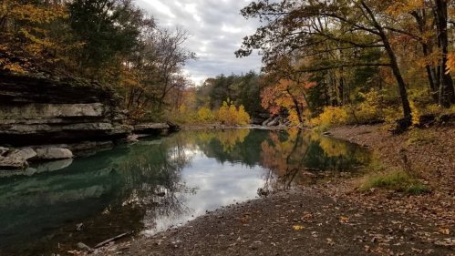 A serene river scene surrounded by autumn foliage and rocky banks, reflecting the cloudy sky above.