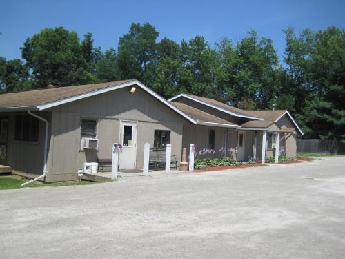 A row of single-story buildings with a gravel path, surrounded by trees and greenery on a sunny day.