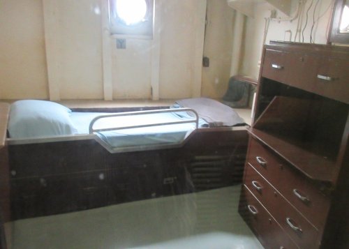 A small, simple bedroom with a bed, dresser, and round window, featuring minimal furnishings and a cozy atmosphere.