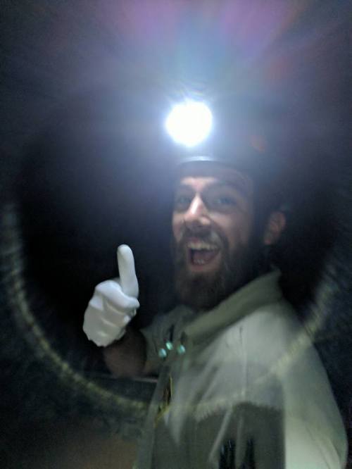 A person in a headlamp and gloves smiles and gives a thumbs up in a dark tunnel.