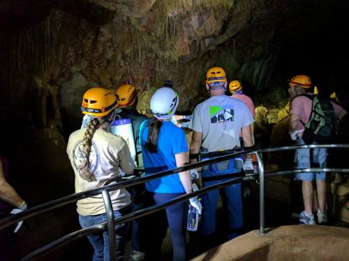 A group of people in helmets exploring a cave, with rock formations illuminated in the background.