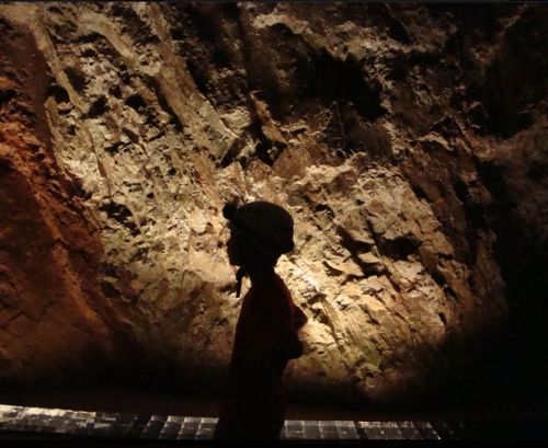 Silhouette of a person wearing a helmet against a rocky, illuminated cave wall.