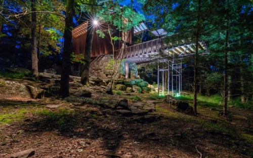 A modern treehouse at night, surrounded by trees and rocks, illuminated by soft green and white lights.