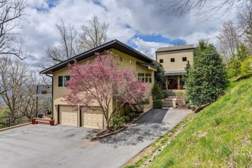 A modern two-story house with a landscaped driveway and blooming trees, set against a cloudy sky and hillside.