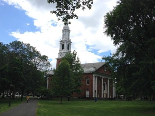 A brick church with a tall clock tower, surrounded by trees and a grassy park under a partly cloudy sky.