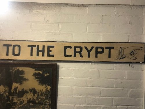 Sign reading "TO THE CRYPT" with an illustration of a pointing hand, mounted on a white brick wall.