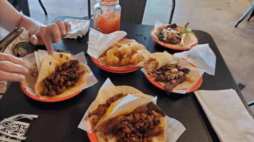 A table with various tacos and snacks, including meat-filled tortillas and a drink in a jar. Hands reaching for food.