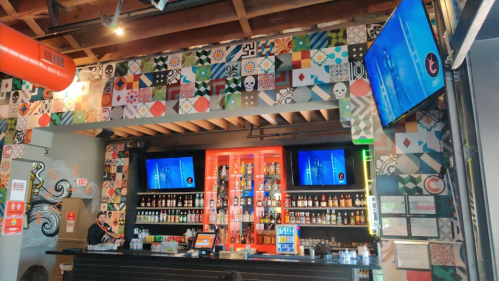 A vibrant bar interior with colorful wall art, a well-stocked shelf, and two screens displaying images.