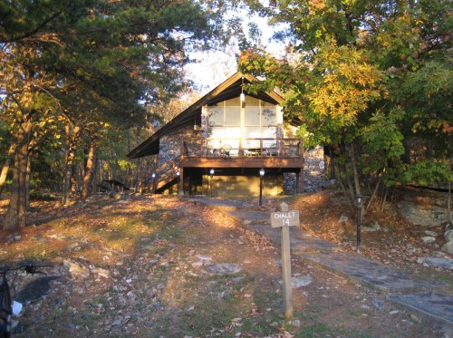 A cozy chalet nestled among trees, featuring a stone facade and large windows, with a sign indicating "Chalet 14."