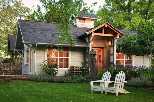 A charming gray house with a porch, surrounded by greenery and flowers, featuring two white Adirondack chairs in the yard.