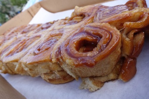 A close-up of a caramel cinnamon roll, drizzled with syrup, resting on a white paper in a cardboard box.