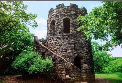 A stone tower with a spiral staircase, surrounded by lush greenery and trees under a partly cloudy sky.