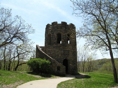 Stone tower with arched windows, surrounded by trees and grass, under a clear blue sky. Pathway leads to the entrance.