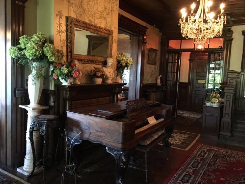 A vintage piano in an elegant room with floral arrangements, a chandelier, and ornate wooden decor.