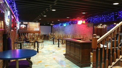 A dimly lit bar interior with colorful lights, empty tables, and chairs stacked on the bar.