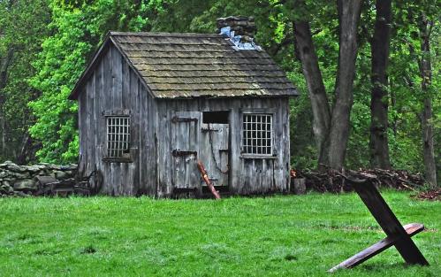 A weathered wooden shed with a stone chimney, surrounded by lush green trees and grass.