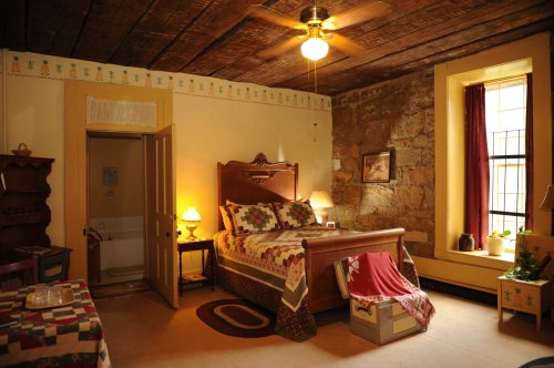 Cozy bedroom with a wooden bed, quilt, stone wall, and warm lighting, featuring a window and decorative furnishings.